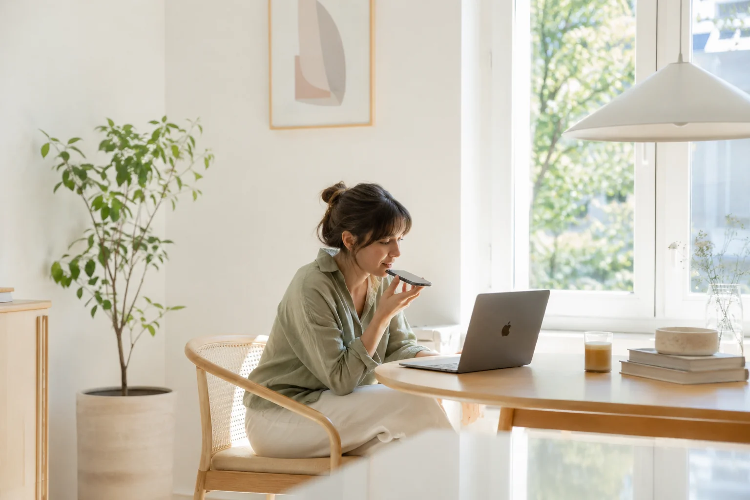 In a sunlit Nordic-style room with houseplants and a closed MacBook nearby, a woman holds her phone close to her lips and speaks softly — turning a quiet moment into captured thoughts.